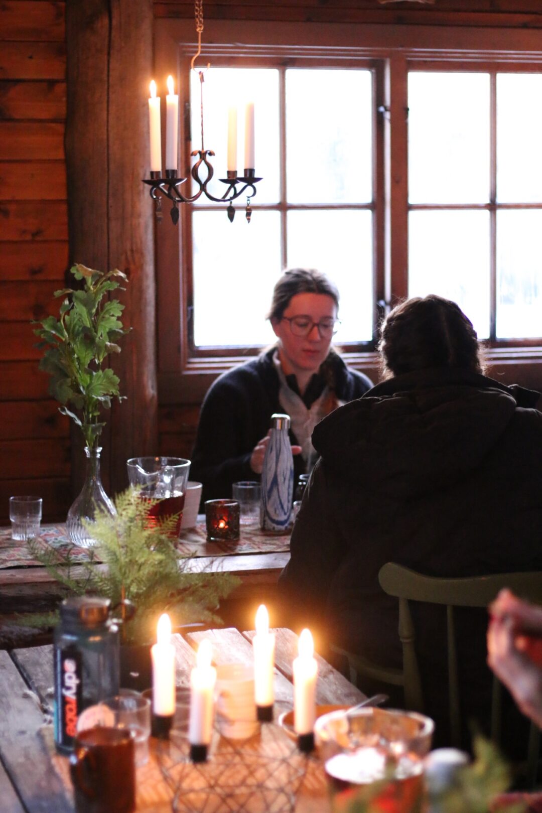 Women dining by candlelight in a wooden cabin, discussing drinks by a window.