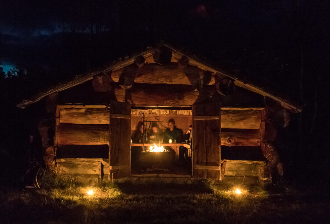 Warmly lit wooden outdoor shelter with a central fire, lit lamps and cosy log walls at night in Sweden