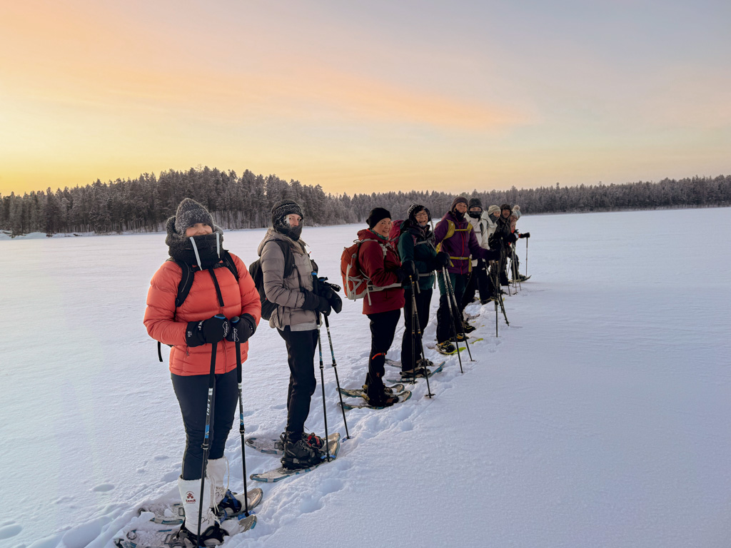 Women snowshoeing on a frozen lake in Finland at sunset, standing in a line with poles.