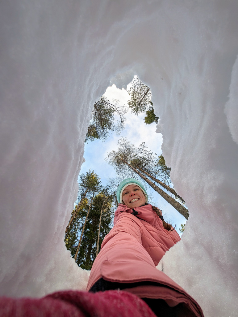 Smiling woman in winter clothing in deep snow, looking up at pine trees through a snowshoe track