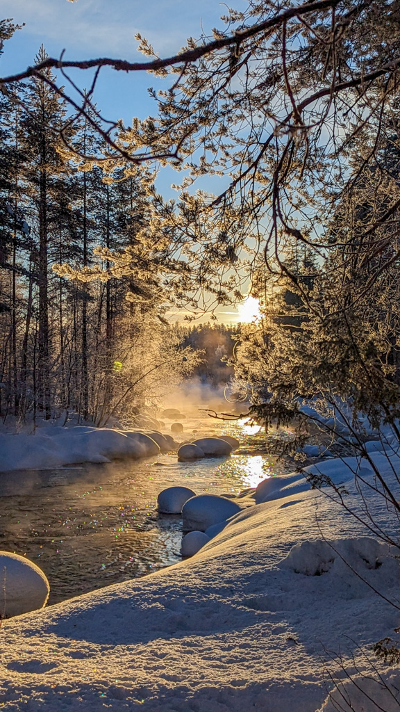 Snow-covered forest with a partly frozen river at sunrise, with snowshoer tracks along the bank