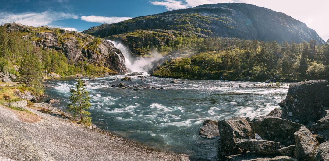 Rushing waterfall into a green fjord along a rocky trail in Norway, surrounded by mountains