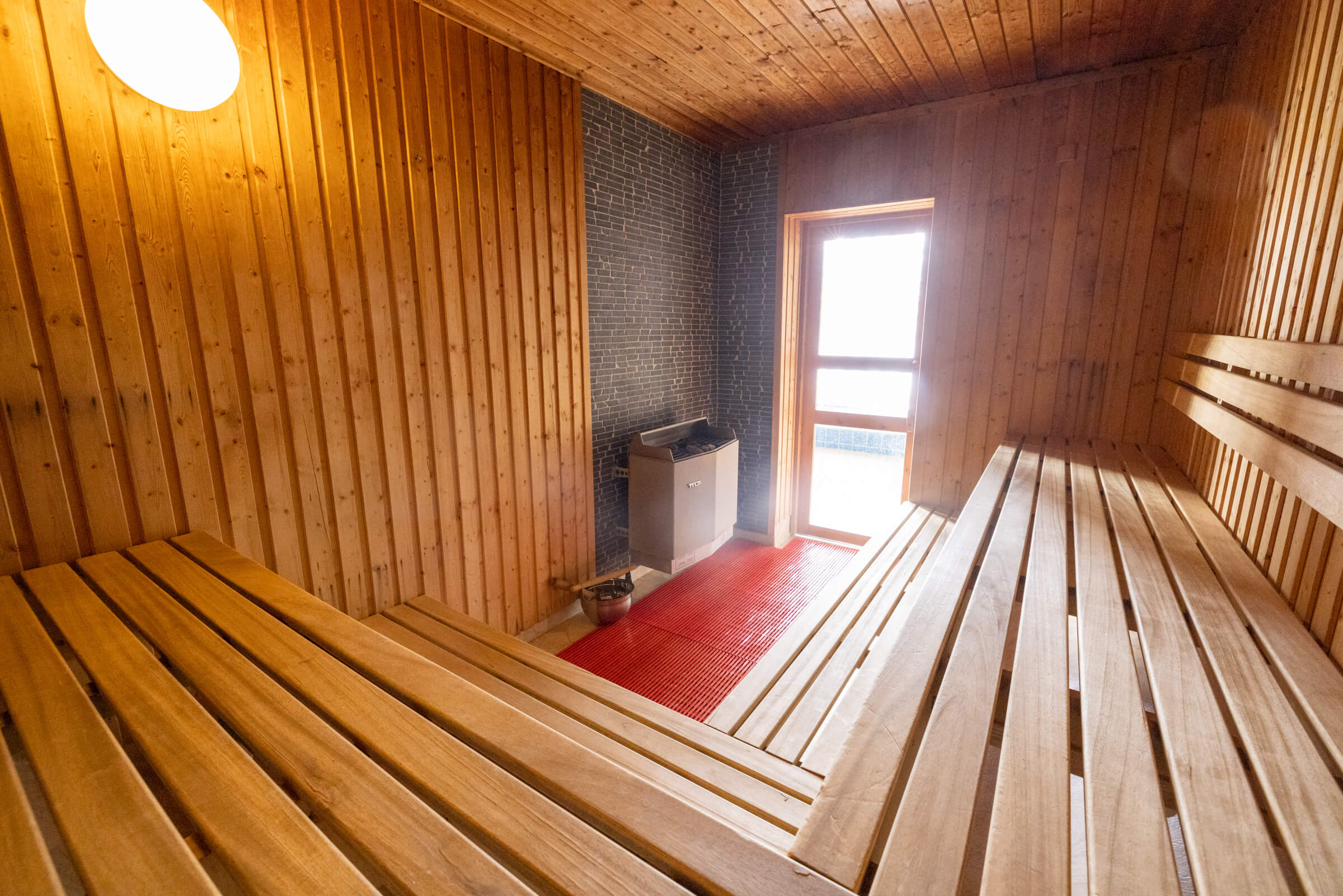 Wooden sauna interior with brick stove, bench seating and window light.
