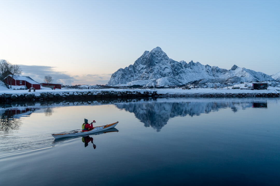 Woman kayaking on calm fjord near snowy mountains and cabins in Lofoten, Norway