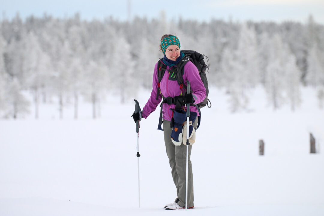 Woman backcountry skiing in a snowy forest, wearing a purple jacket with skis and poles.