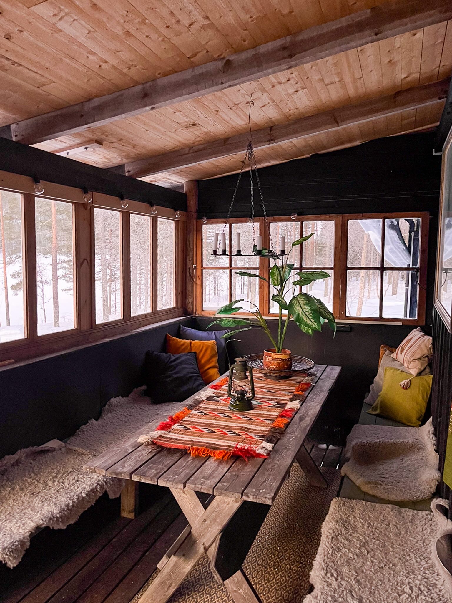 Wooden cabin interior with table and hanging lamps, overlooking snowy forest in Sweden