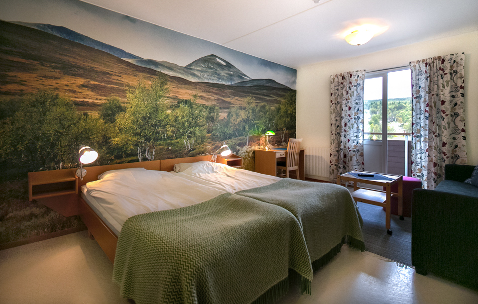 Hotel bedroom with double bed, bedside lamps, and mountain landscape mural on the wall, plus a window.
