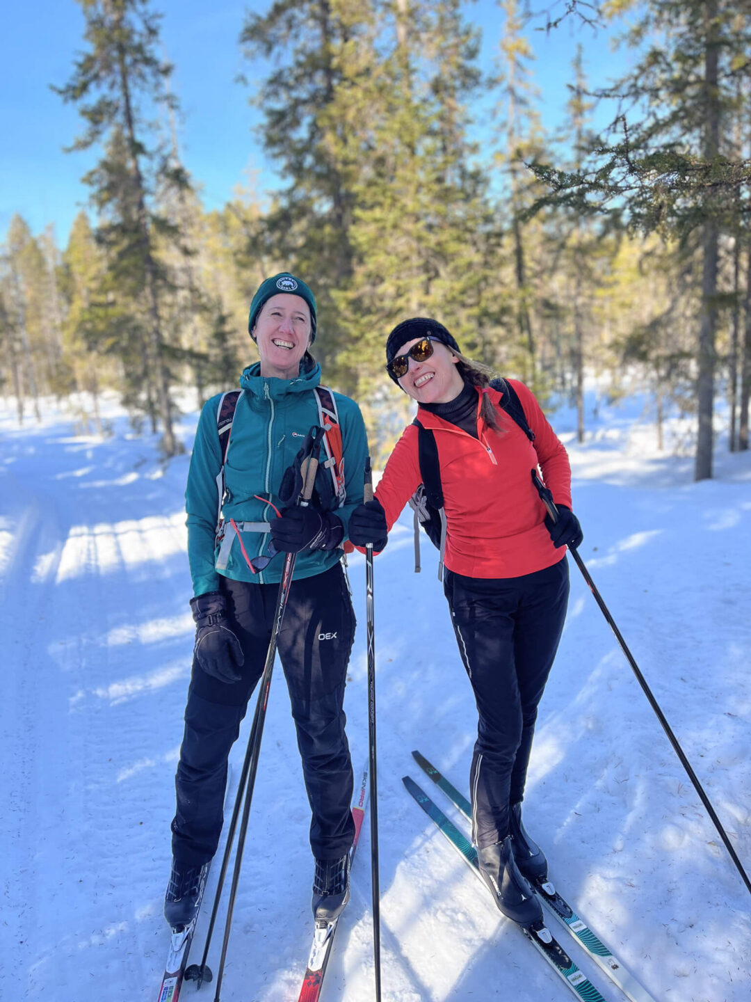 Two cross-country skiers smiling on skis in a snowy Finnish forest trail under blue sky