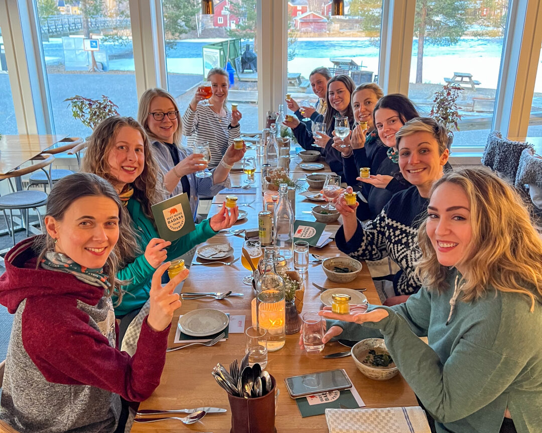 Friends in a cosy Swedish Lapland restaurant toast with drinks by a window, snow and dog sledding scenery outside