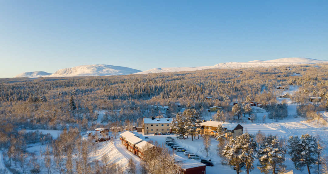 Snowy Swedish countryside aerial view with a winter lodge, pine forest and distant hills under clear blue sky