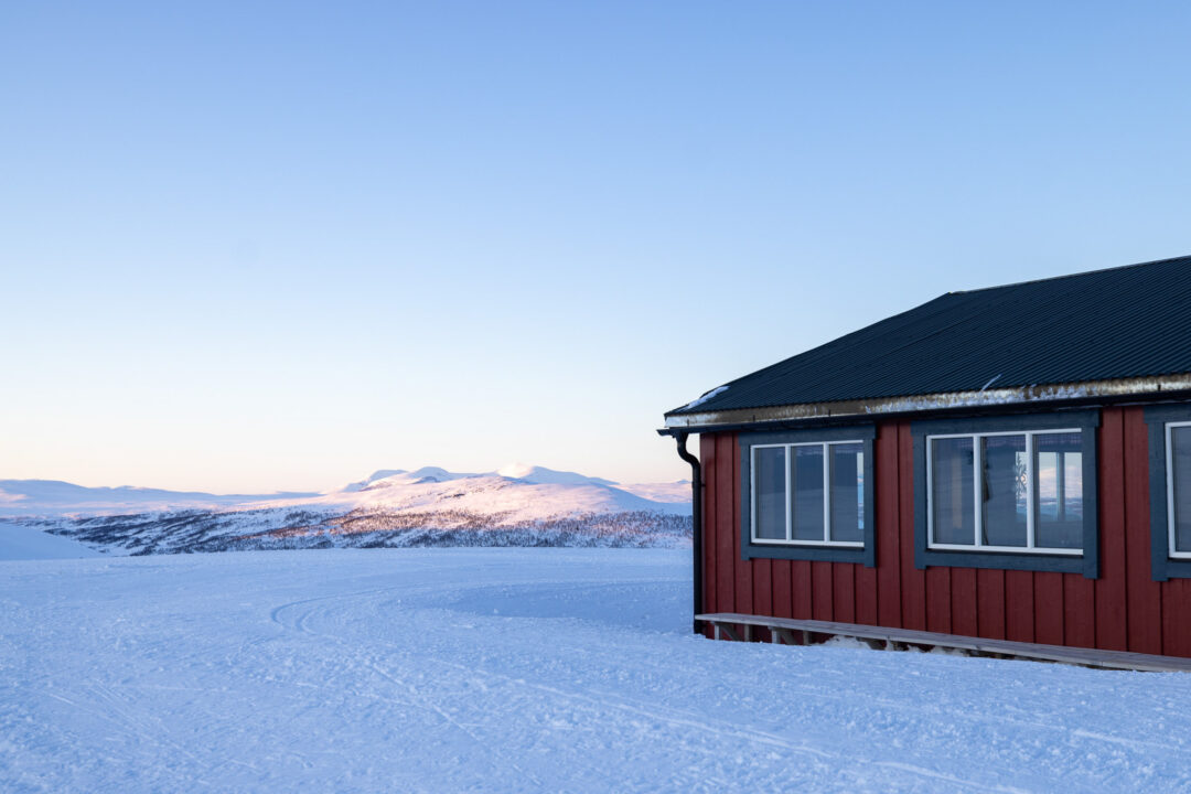Red wooden cabin on a snowy Swedish winter landscape at dusk with distant mountains on the horizon