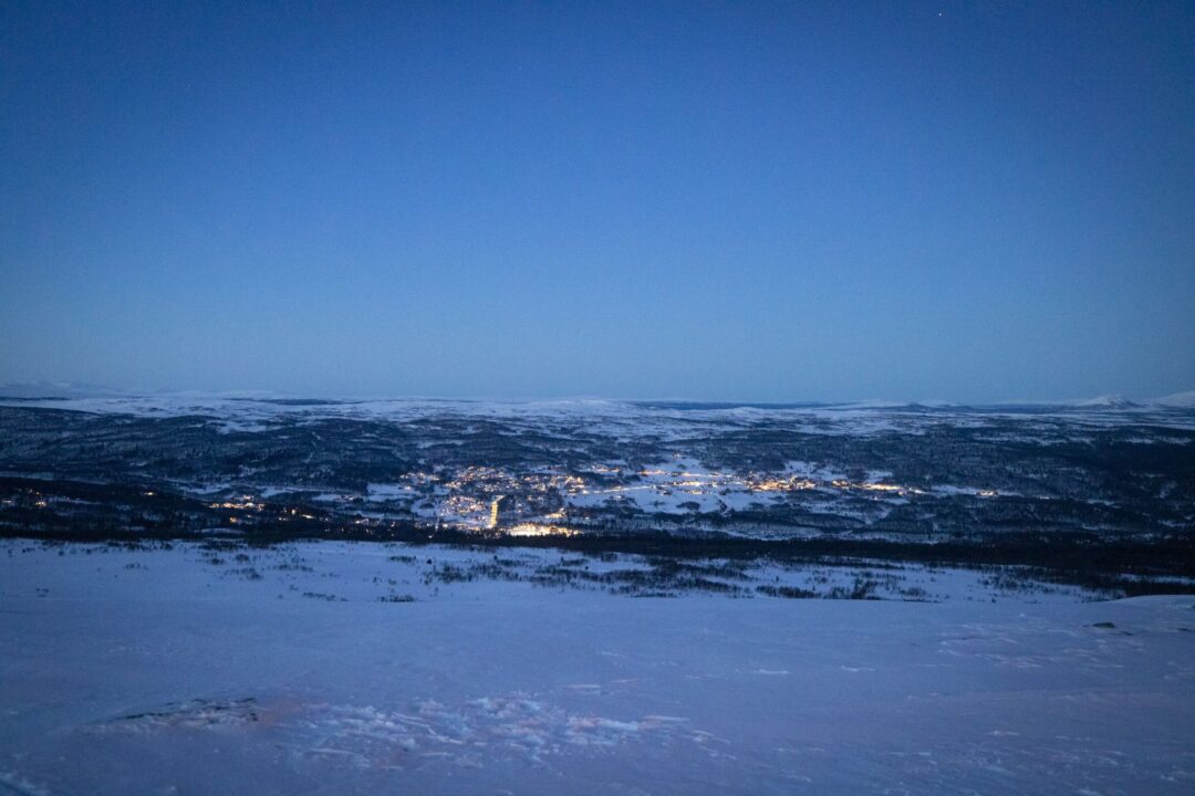 Snowy landscape at night with a valley town lit up in the distance and clear winter sky