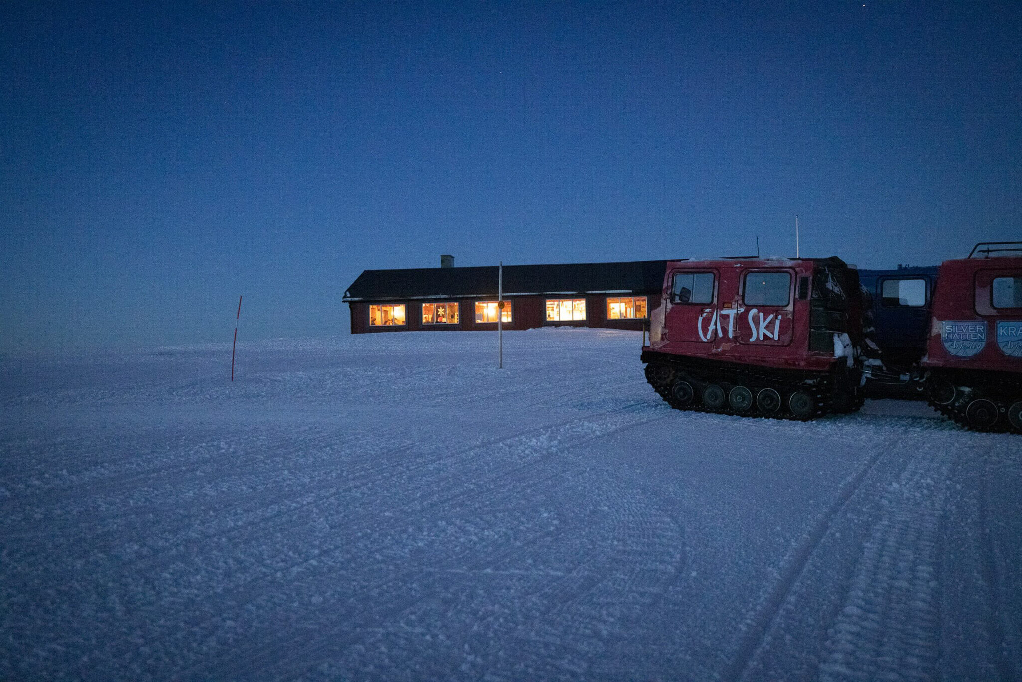 Snowcat and lit ski lodge on snow tracks at dusk in Sweden, with “CAT SKI” on the vehicle