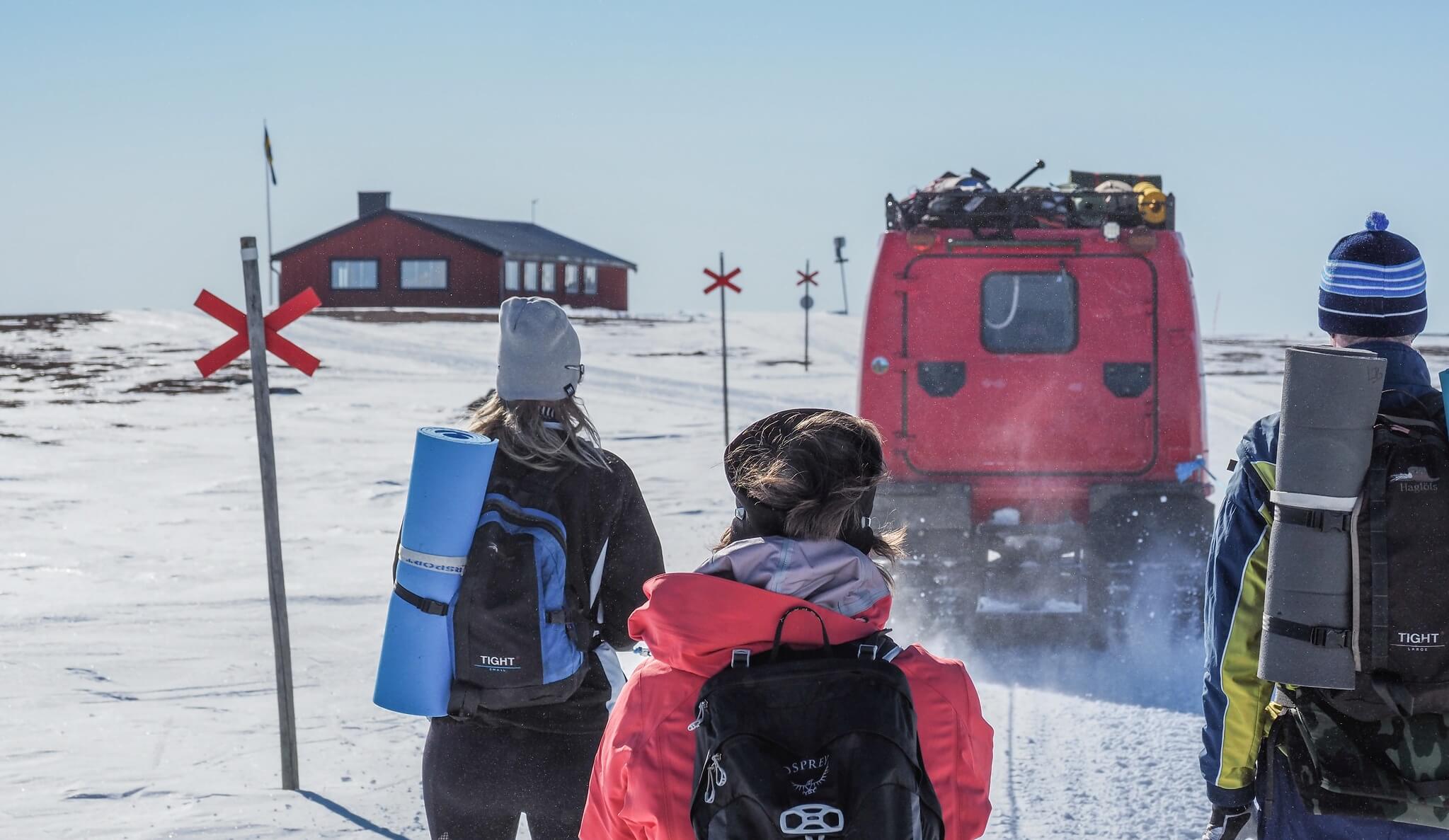 Women in winter clothing with yoga mats and backpacks walk past a red snow vehicle in snowy Sweden