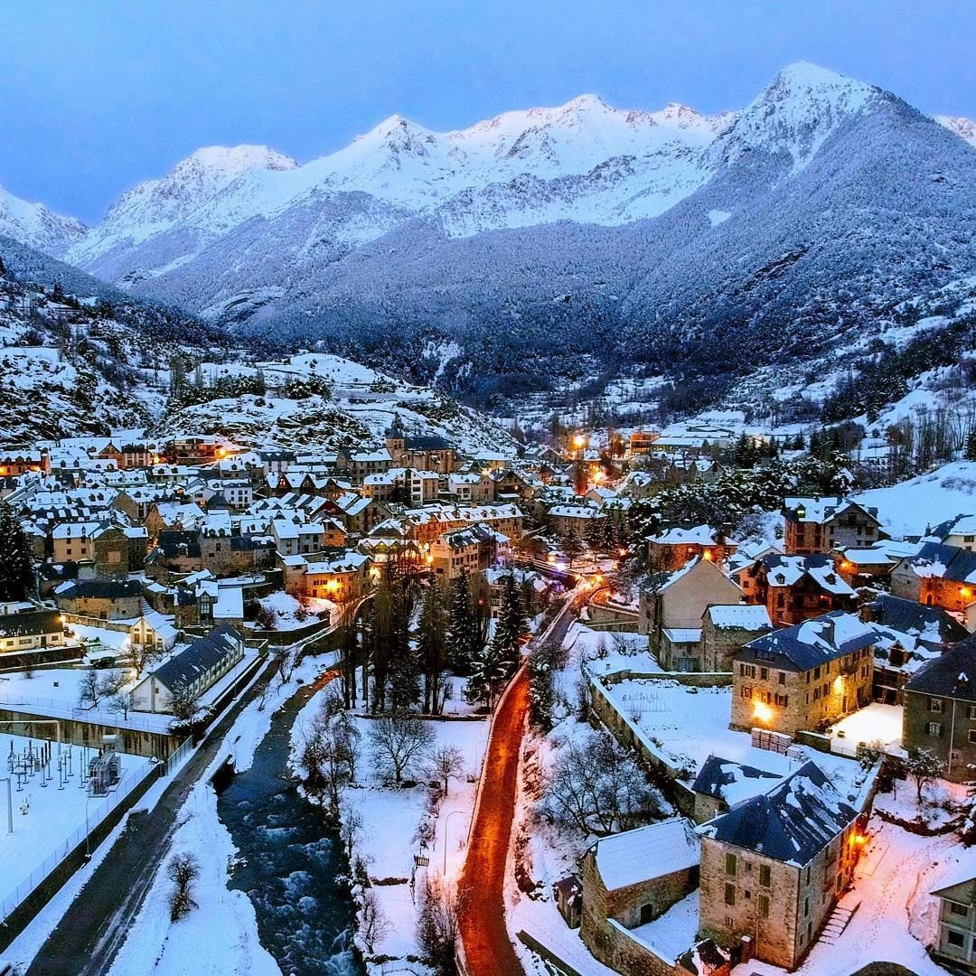 Snowy Pyrenees village at dusk with winter hiking tracks, river and mountain peaks in view