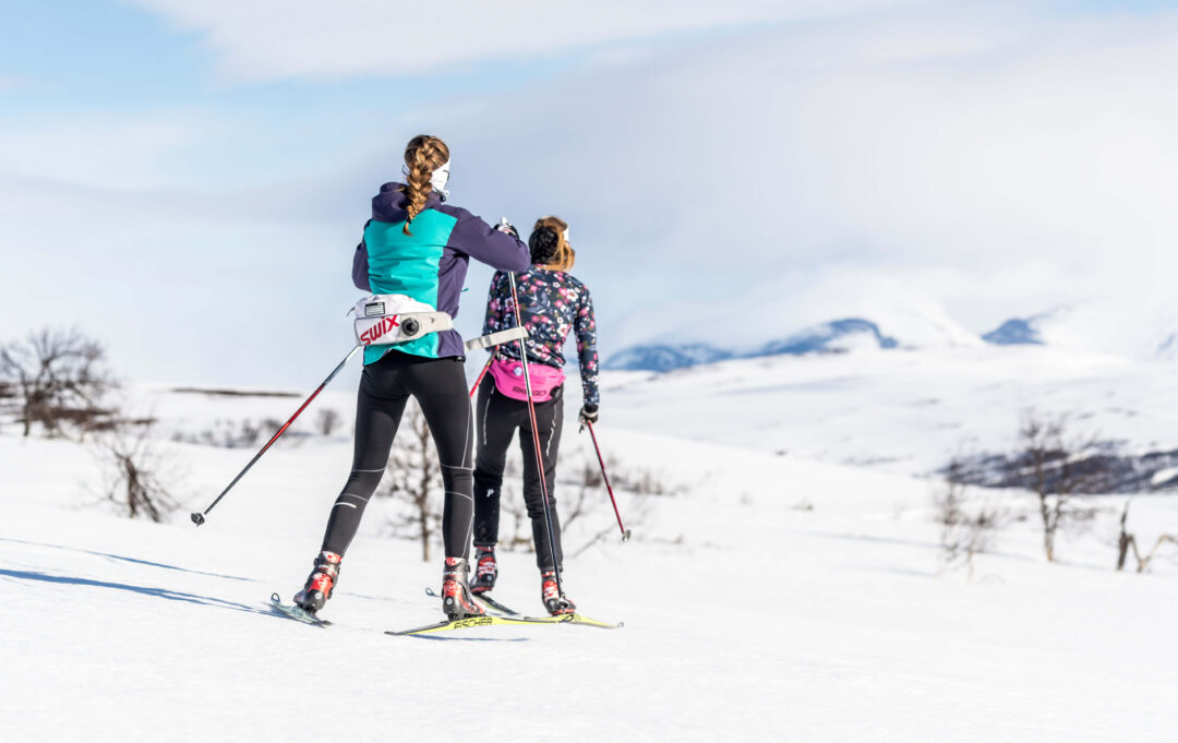 Two women skate skiing in snowy Sweden, with skis and poles; “SWIX” logo on a hat.