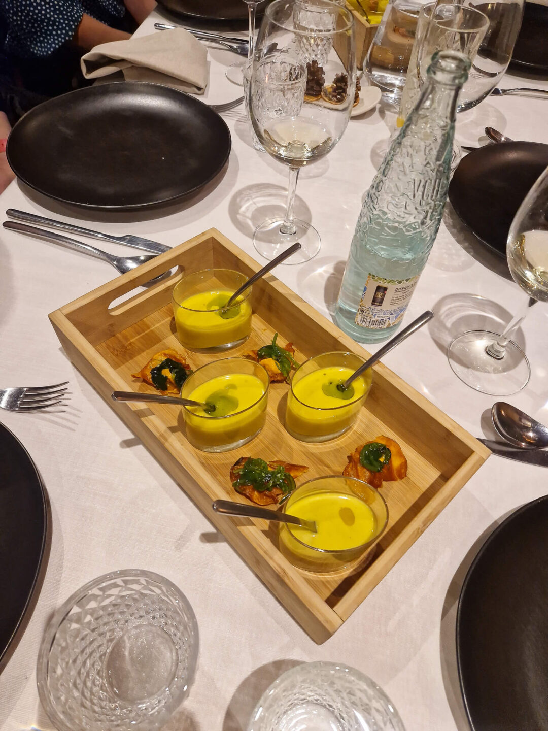 Dining table with yellow aperitifs in glasses and a bottle, set with plates and cutlery indoors