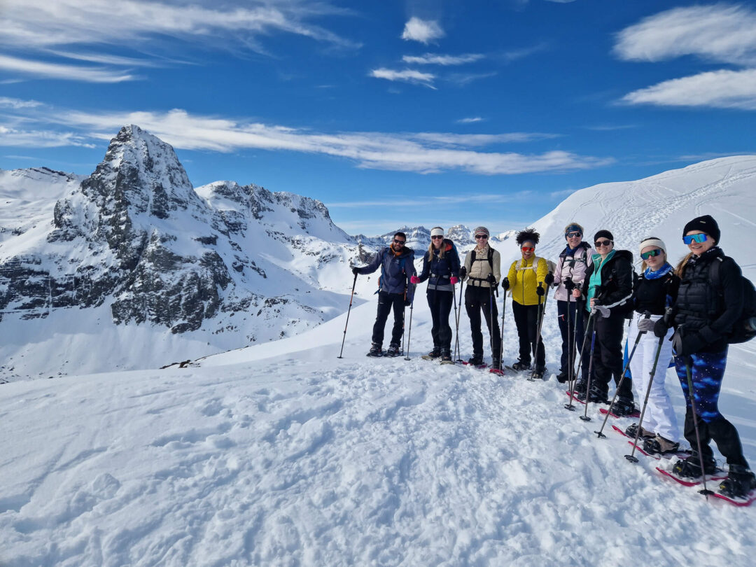 Snowshoeing group on a snowy Pyrenees ridge with trekking poles and mountain peaks under blue sky