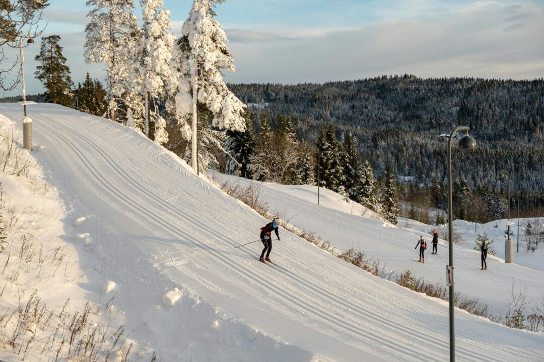 Cross-country skiers on a groomed snow trail through a winter forest and hills.