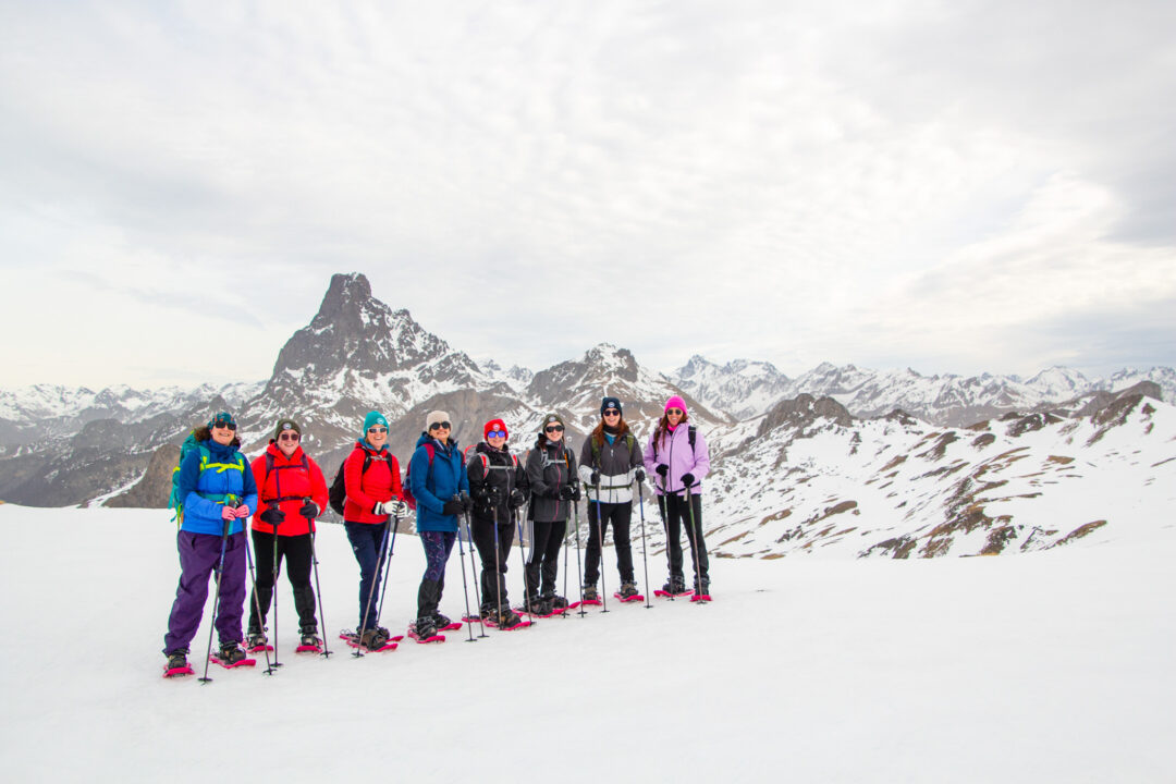 Women snowshoeing on a snowy mountain ridge in winter, standing with trekking poles and snow gear against peaks