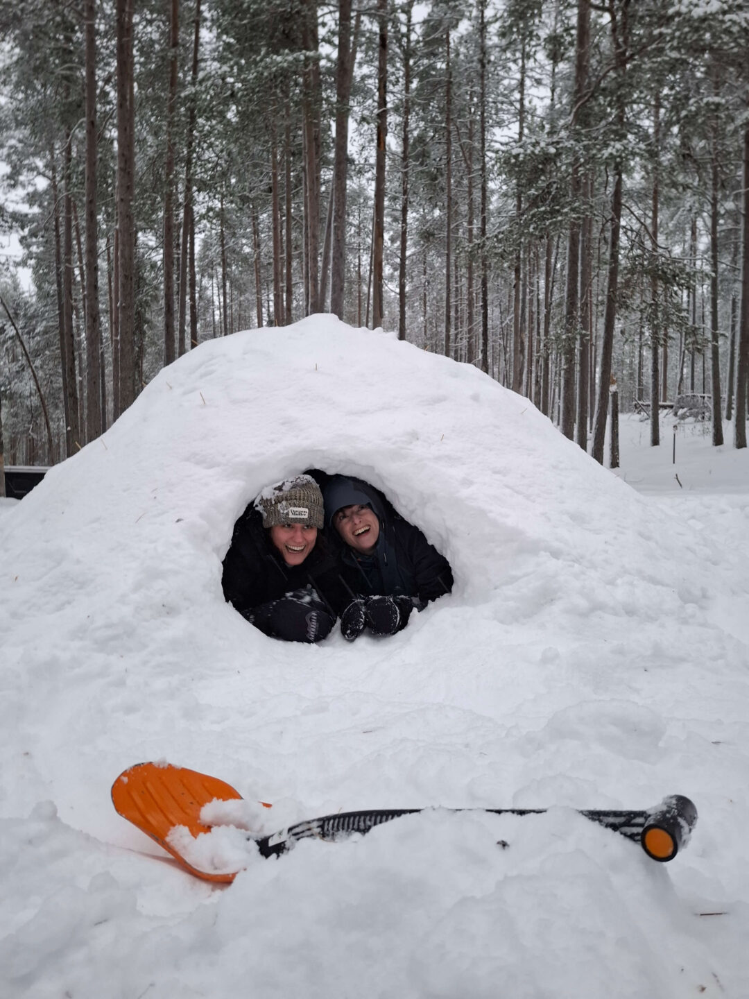 Two snowshoers smiling in a snowy shelter cave in a Finland pine forest, snowshoes visible