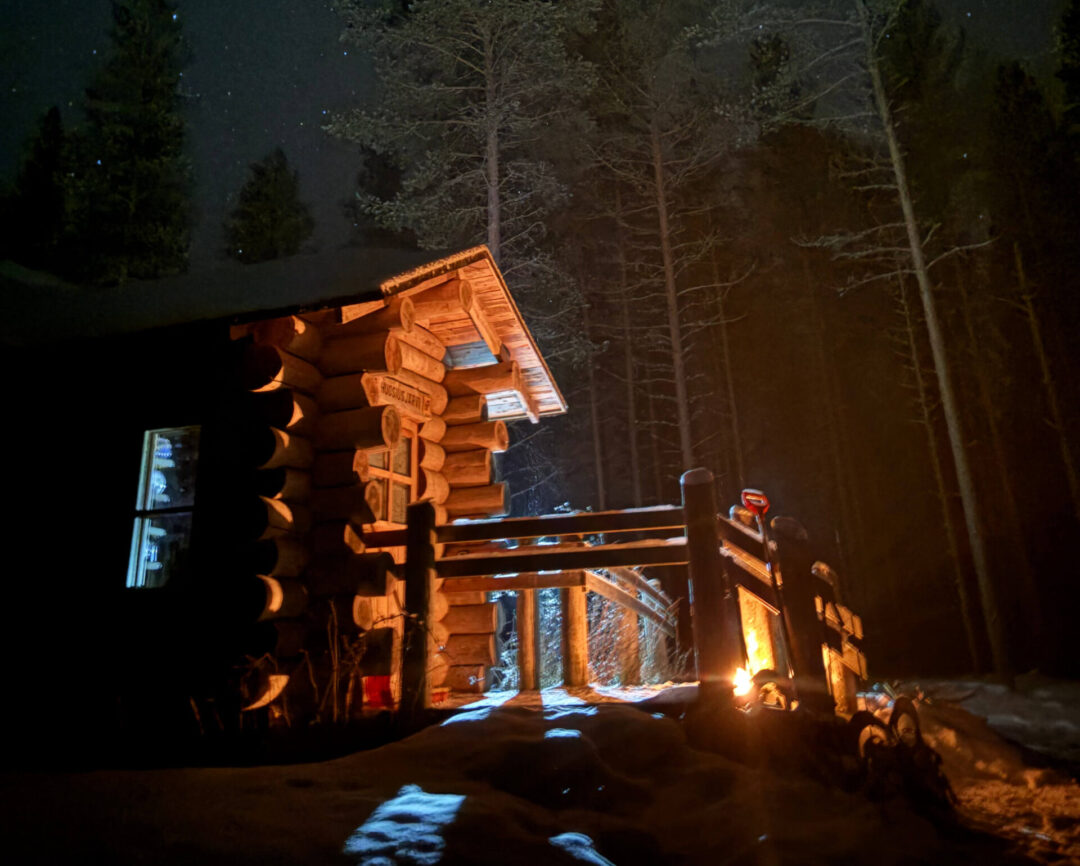 Warmly lit wooden cabin in a snowy Finnish forest at night, with snowshoe tracks and darkness beyond