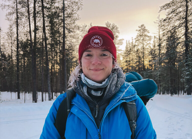 Woman in blue jacket snowshoeing through a snowy Finnish pine forest at sunset, wearing a red beanie.