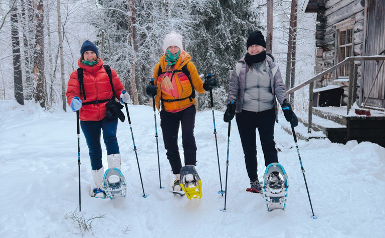 Three people snowshoeing through Finland’s snowy forest near a wooden cabin in winter.