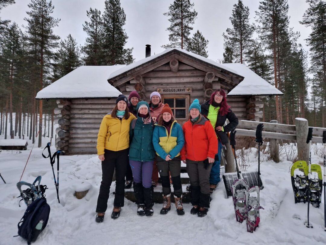 Women posing outside a snowy log cabin in Finland; skis and sign “HUOSJARVI” by the door