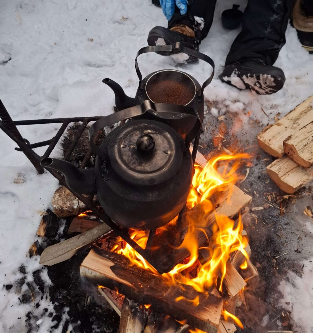 Kettle boiling over an open campfire in snowy outdoor conditions, with a woman’s boots nearby