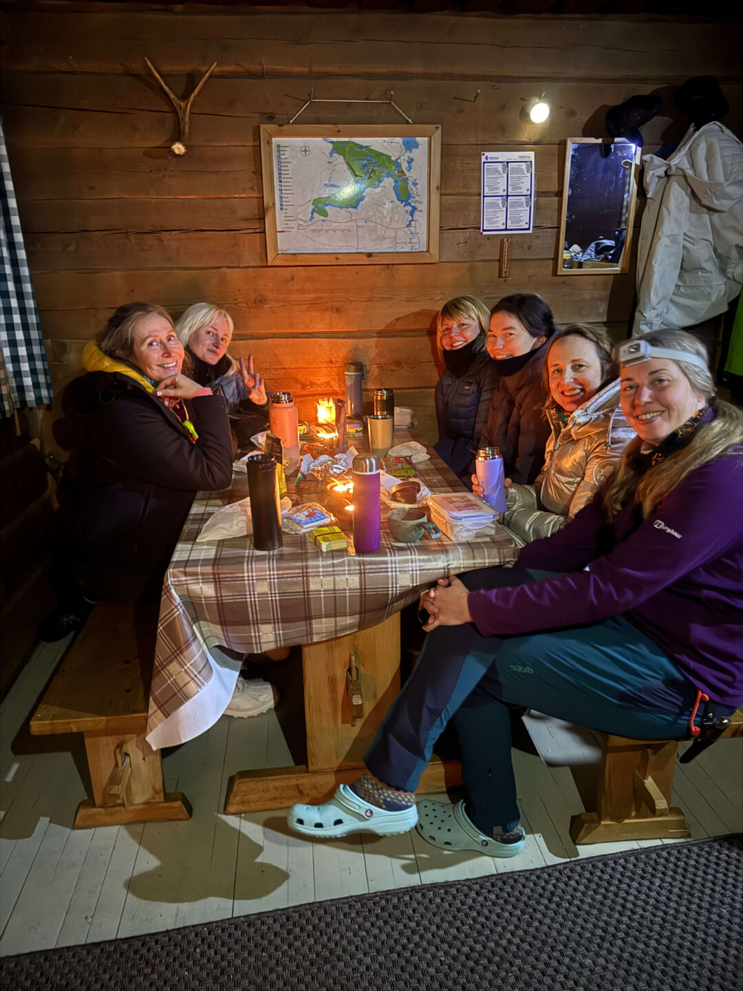 Women sit around a lit table in a wooden cabin, with a wall map and notices behind them