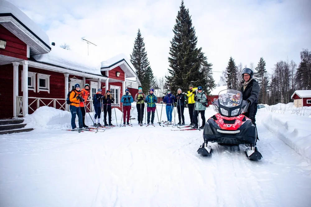 Cross-country skiers lined up in Finland near red cabins on a snowy trail, ready to ski