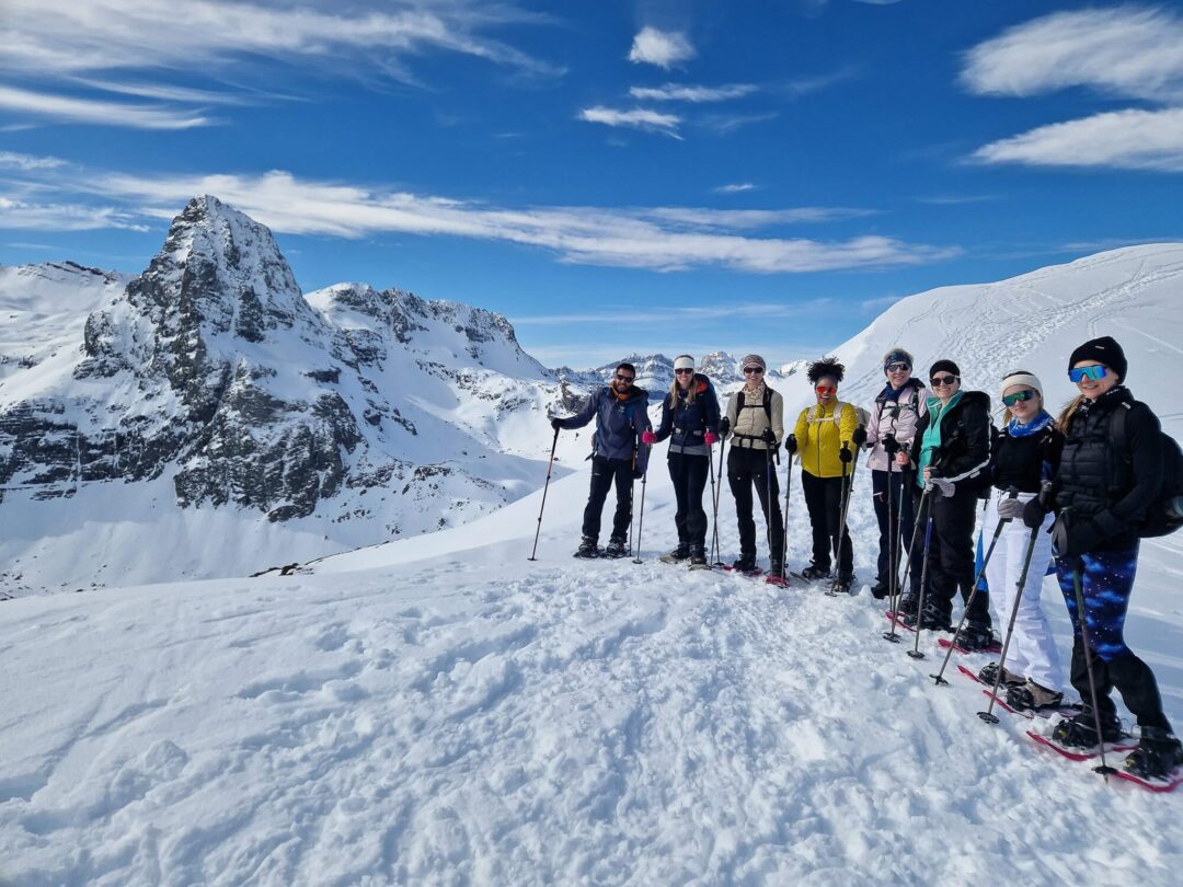 Women snowshoe hikers on a snowy mountain ridge in the Pyrenees, posing in winter sunshine with poles