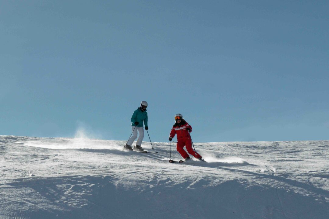 Two women skiing downhill on a snowy slope under a clear blue sky