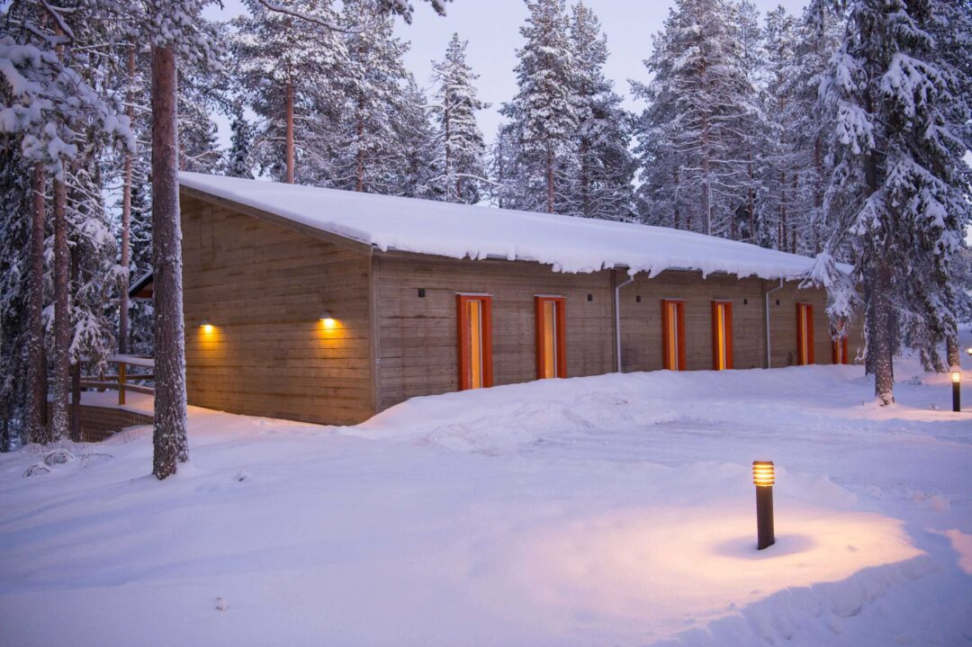 Snowy Swedish Lapland cabin in a winter forest at dusk, with warm lights glowing in the terrace snow.