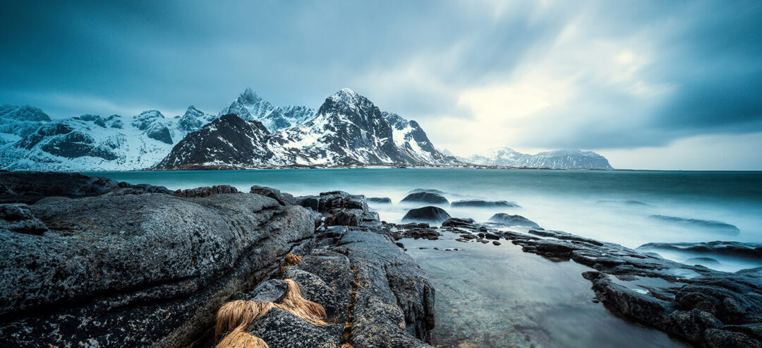 Snowy rocky coastline in Lofoten, Norway, with winter sea and mountain peaks under a cloudy sky.