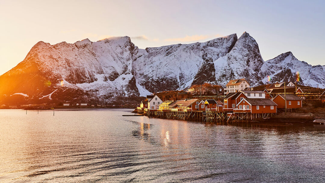 Snowy Lofoten mountains and waterfront village with red houses at sunrise, reflecting in the fjord.