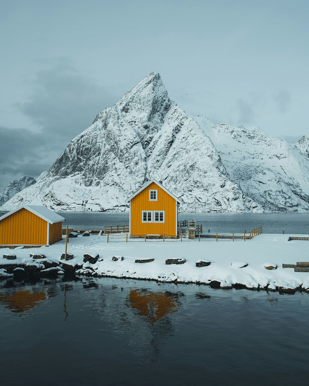 Snow-covered yellow fishing cabins by the fjord with steep mountains in Lofoten, Norway
