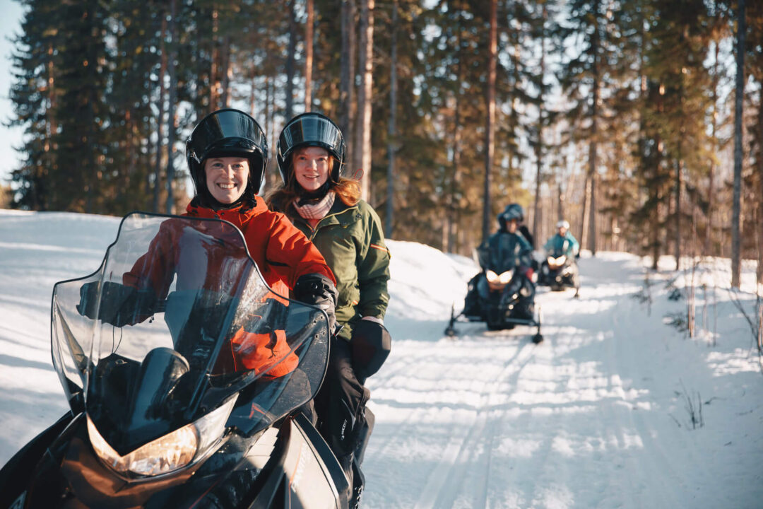 Two people on snowmobiles smiling in a snowy Lapland forest during a winter multi-activity adventure