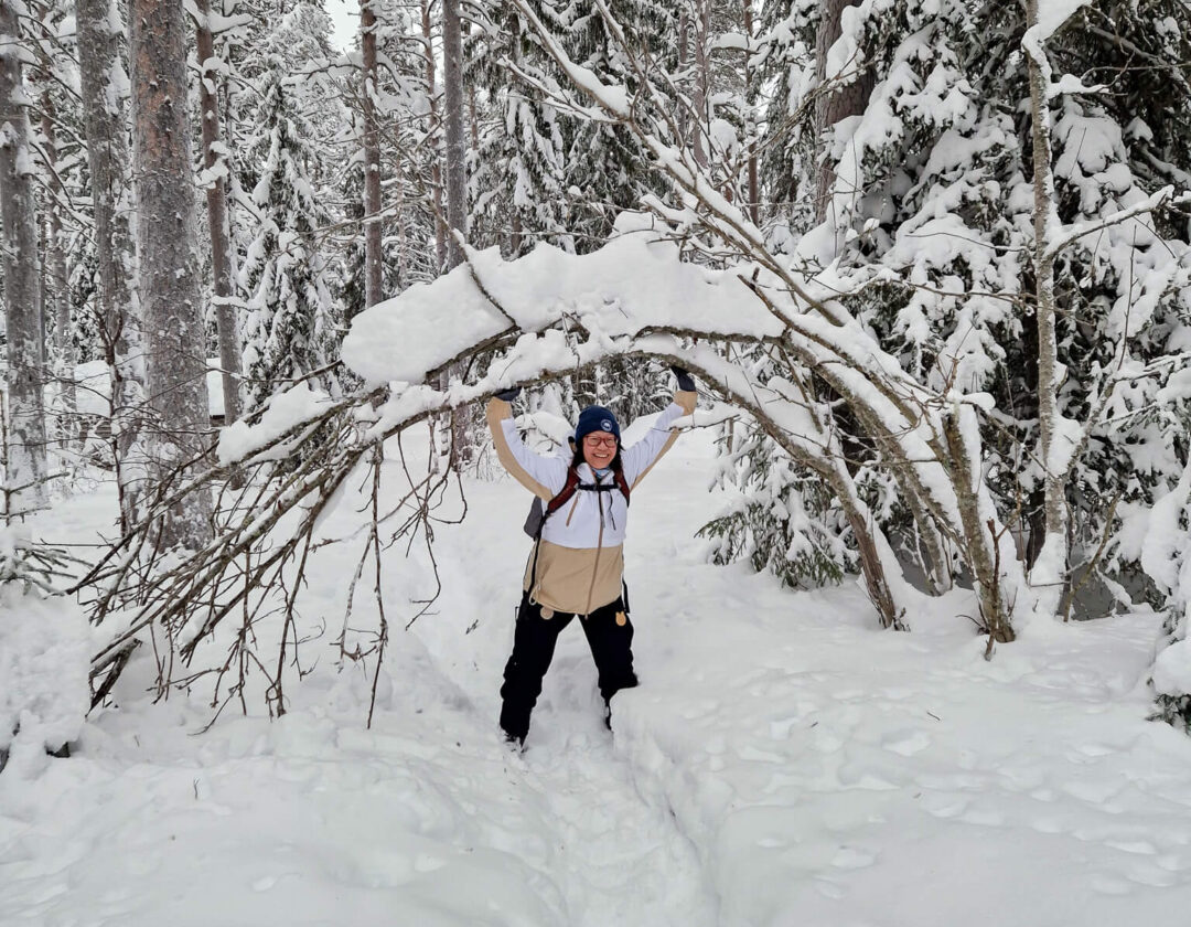 Snowshoeing woman standing in a snowy pine forest, arms raised amid fresh Finland-style winter snowfall
