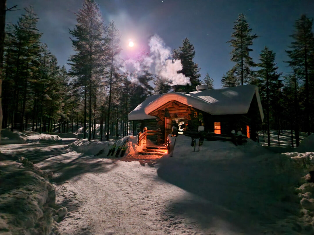 Warm log cabin in snowy Finland forest at dusk, misty smoke rising beside a snowshoe trail