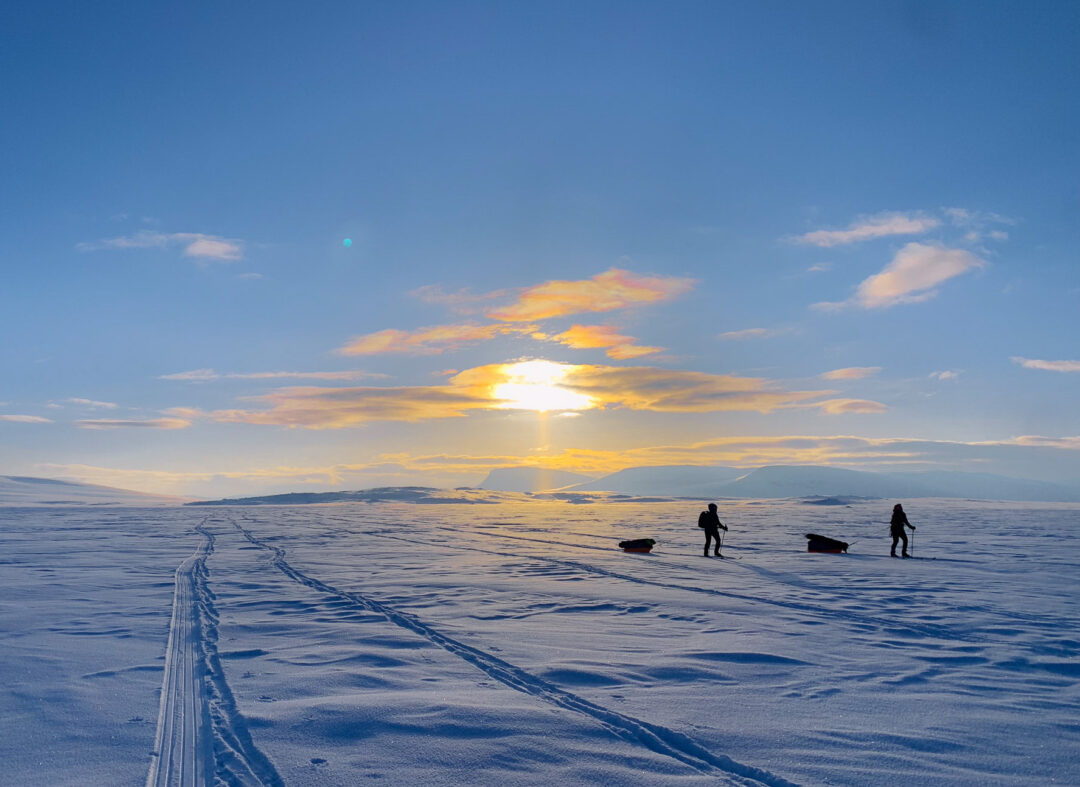 Ski tracks across snowy Arctic tundra at sunrise, with lone skiers heading towards distant huts