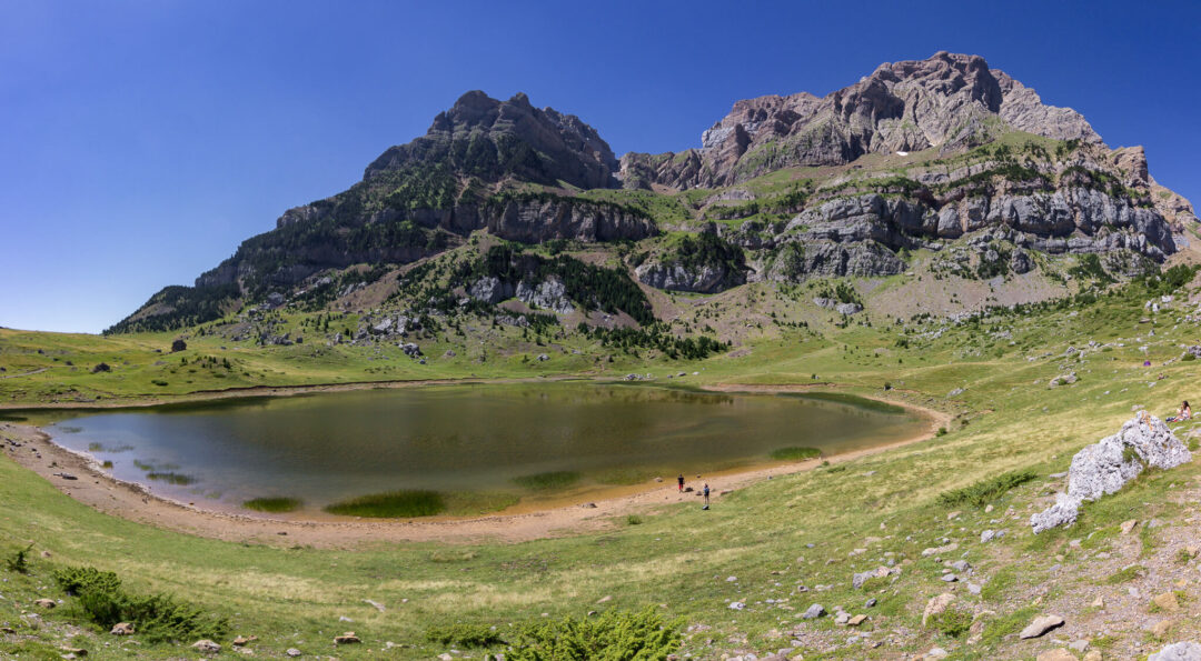 Hikers by a mountain lake in a green valley with rugged peaks and a winding trail