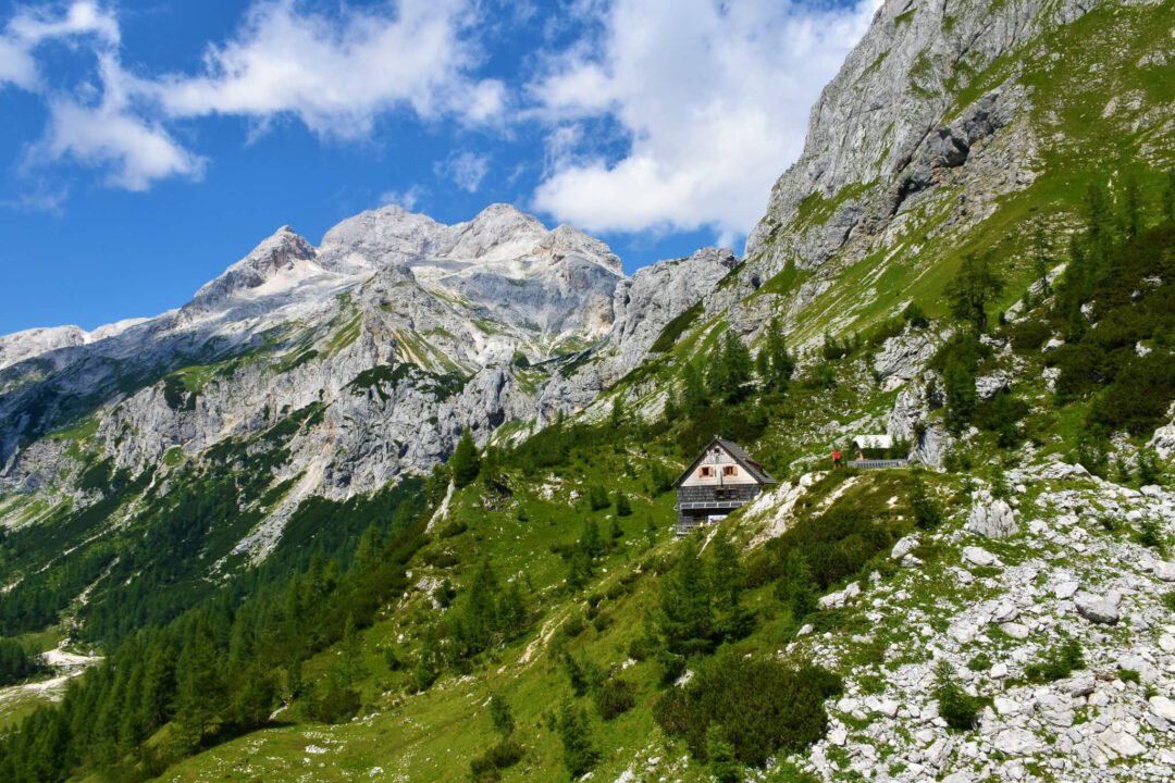 Hut-to-hut hiking route in Slovenia: mountain hut on a green alpine valley with rocky peaks and blue sky
