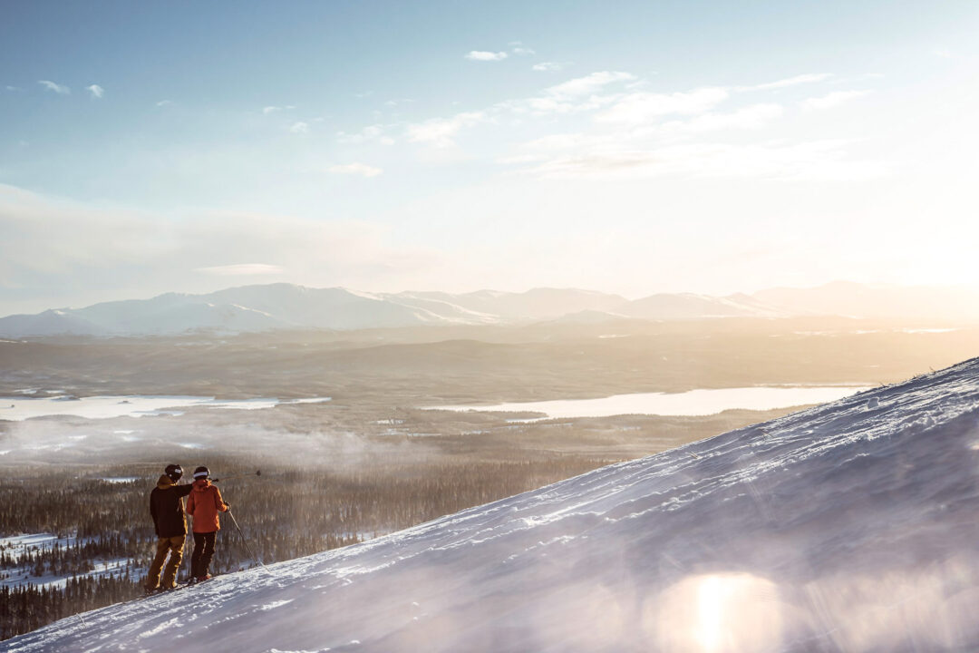 Beginner downhill skiers on a snowy Swedish mountain slope at sunset, overlooking ski runs and valleys
