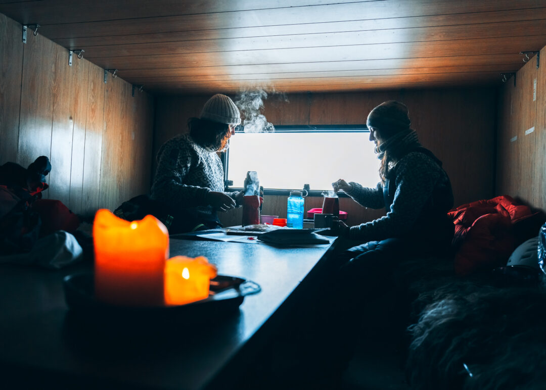 Two snowmobilers in a wooden Svalbard cabin warm up by a table before heading out over snowy ice.