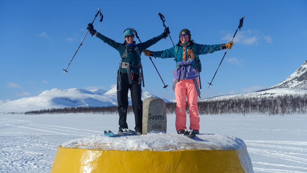 Two cross-country skiers celebrate atop a yellow marker labelled “Suomi 1996” in snowy Arctic landscape