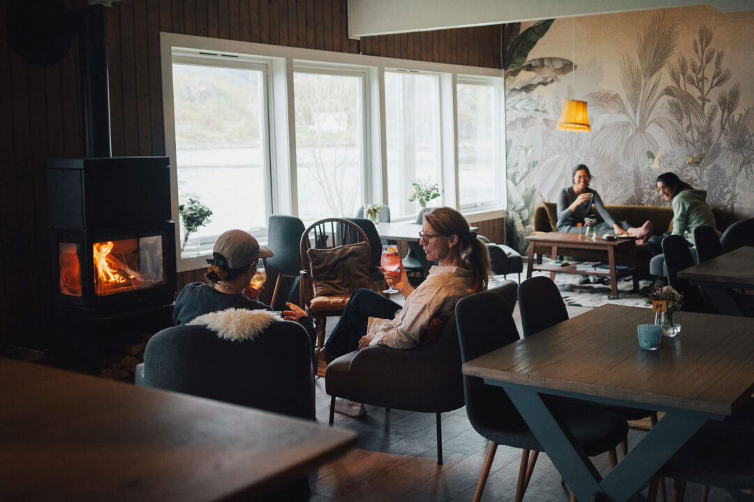 People relaxing around a fireplace in a cosy Lofoten lodge dining room with snowy views through windows