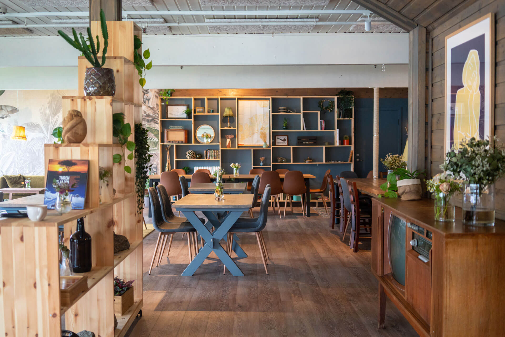 Cafe dining area with wooden tables, blue chairs, and potted plants inside a light-filled room
