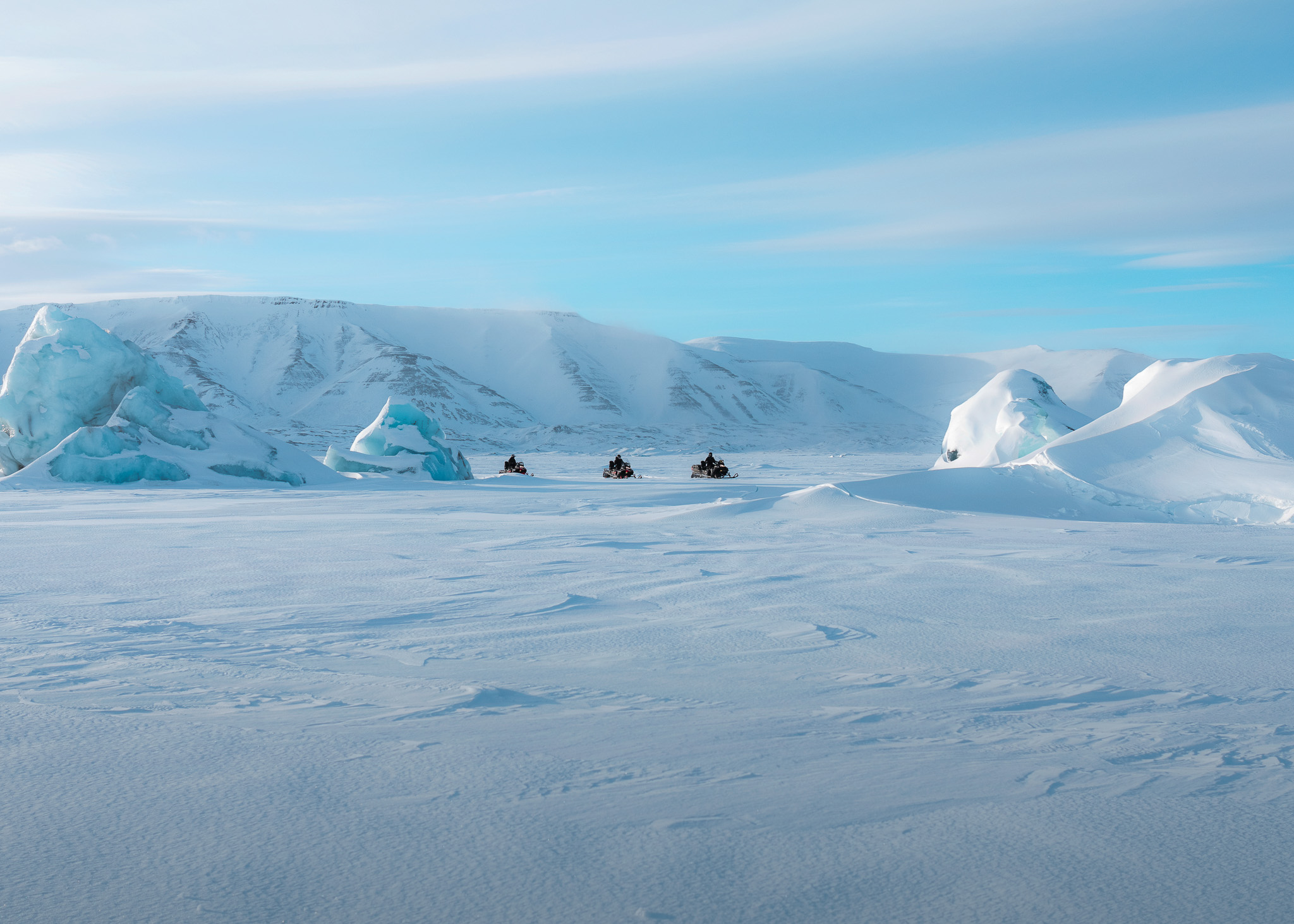 Snowmobiles ride across Svalbard snowfields toward icy mountains under a clear blue sky